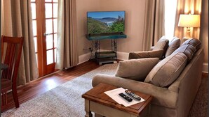 A living room with a flat-screen TV on a stand, a sofa, a wooden coffee table with remotes, and a chair.