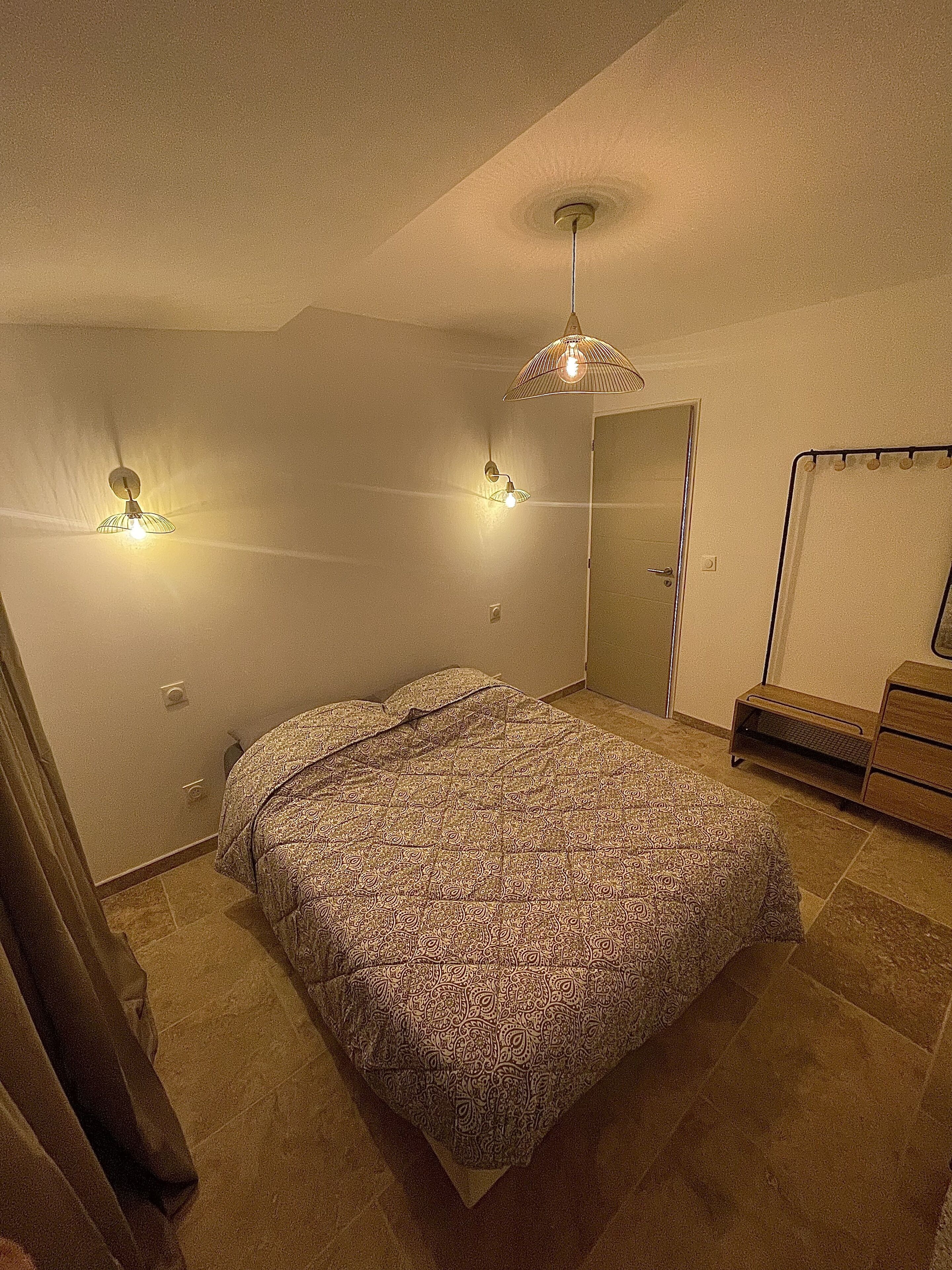 A bedroom with a bed, two wall-mounted lamps, a ceiling fan, a door, and a small wooden dresser.