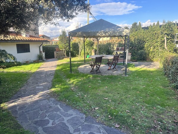 A patio with a table and chairs under a gazebo, surrounded by greenery and a stone pathway.