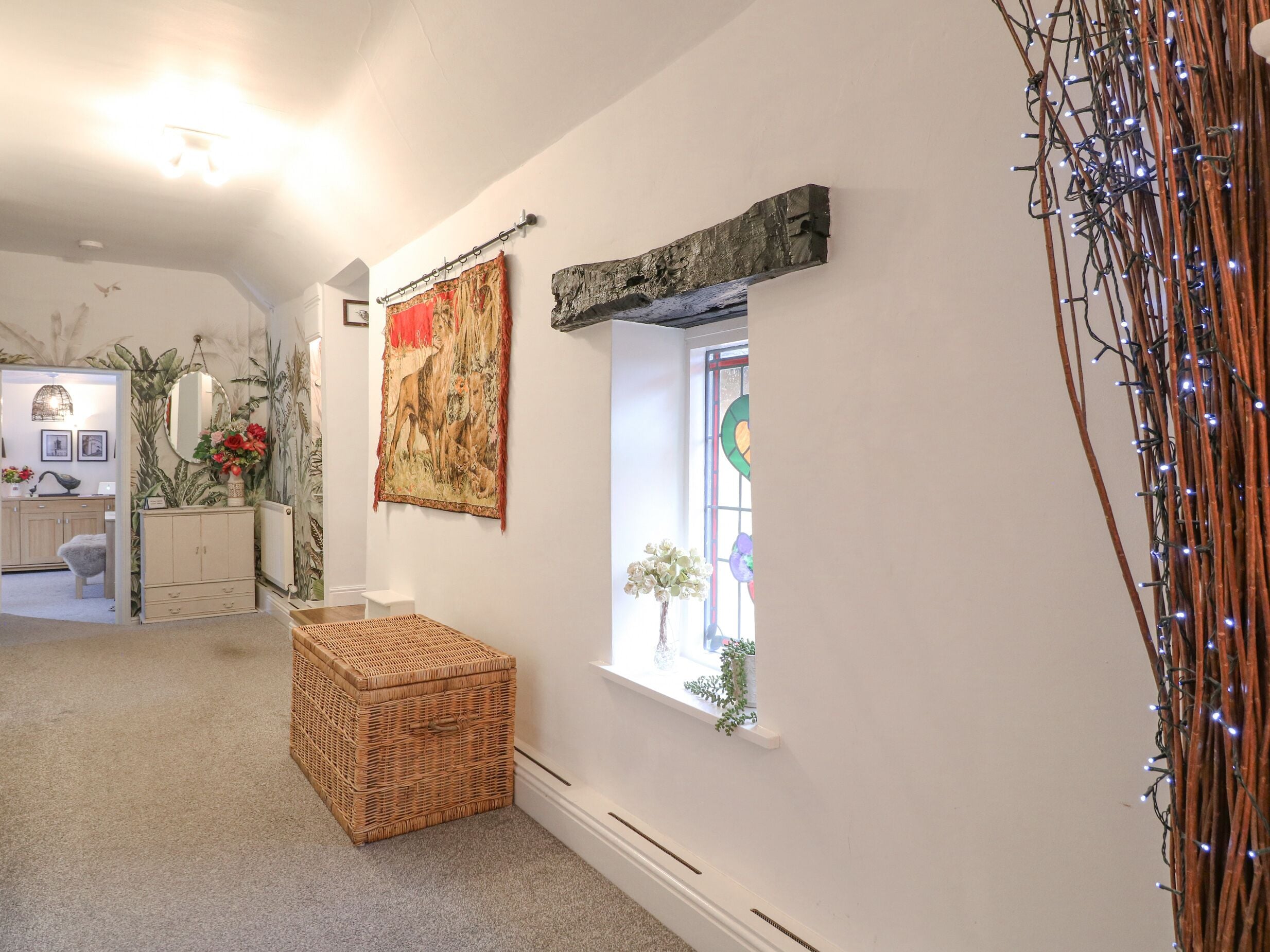 A room with a large tapestry, a small window with a view, a wicker chest, and a decorative light string.