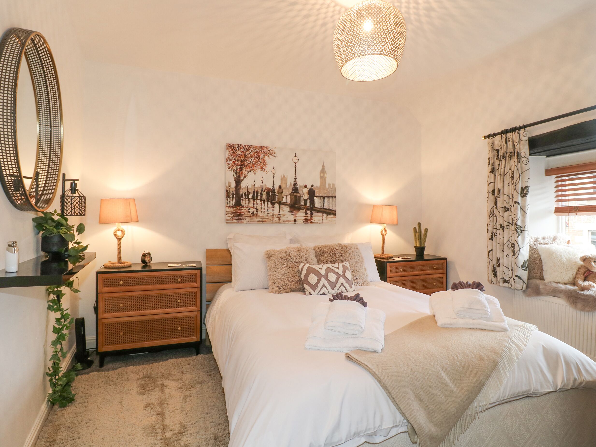 A neatly arranged bedroom with a large bed, two bedside tables with lamps, a wall-mounted mirror, and a cityscape painting.