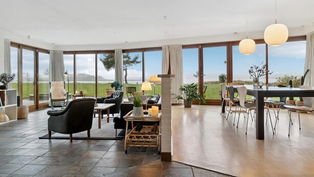 Un vaste espace de vie doté d’une grande fenêtre offrant une vue sur l’extérieur. On y trouve plusieurs options d’assises, notamment un canapé, des fauteuils et une table à manger avec des chaises.