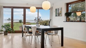 Un espace repas comprenant une table et des chaises, de grandes fenêtres donnant sur l’extérieur et des luminaires suspendus.