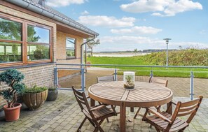 Une terrasse avec une table ronde en bois et des chaises, un pot de fleurs et une vue sur une rivière et des champs.