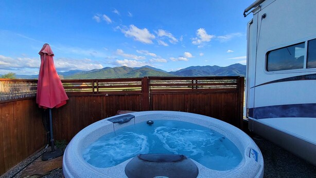 A hot tub with a view of mountains, a wooden fence, and a red umbrella. - 4 units includes two cottages and two trailers with king and queen beds, kitchens, and dining areas. (Talent)