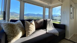 A modern interior with a sofa, large windows, and a view of mountains. - 2 separate RV’s with queen-sized beds, dining area, and full kitchen. (Talent)