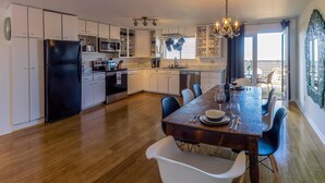 Une cuisine moderne avec des meubles blancs, un réfrigérateur noir, des appareils en acier inoxydable et un coin repas comprenant une table et des chaises en bois.