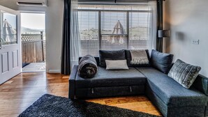 Una sala moderna con un sofá seccional gris, una alfombra negra y vistas a un paisaje urbano a través de ventanas amplias con persianas.