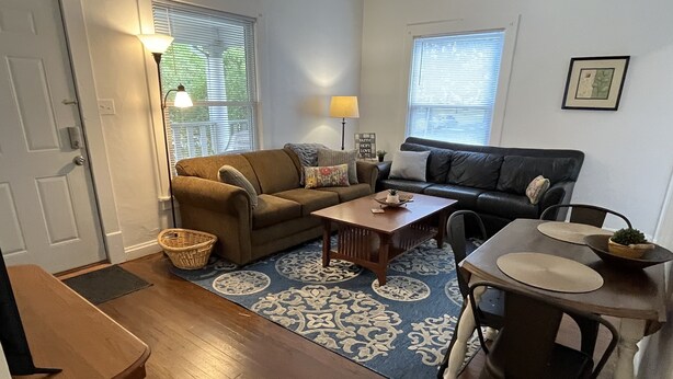 Un salon avec un canapé marron, une table basse en bois et une table à manger avec des chaises. Il y a deux fenêtres avec des stores, une lampe sur la table basse et un tableau encadré au mur.