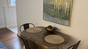 Un coin repas avec une table en bois, quatre chaises, deux sets de table et un saladier rempli de fruits. Un tableau est accroché au mur, au-dessus de la table.