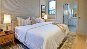 Un dormitorio con una cama grande, dos lámparas de noche, una ventana con persianas, una puerta que da al baño y un ventilador de techo.