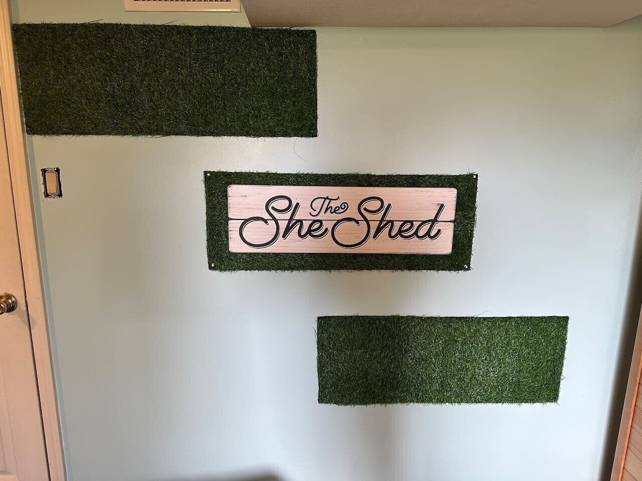 Una puerta con un letrero que dice 'The Shed' en una pared con parches de césped artificial verde.