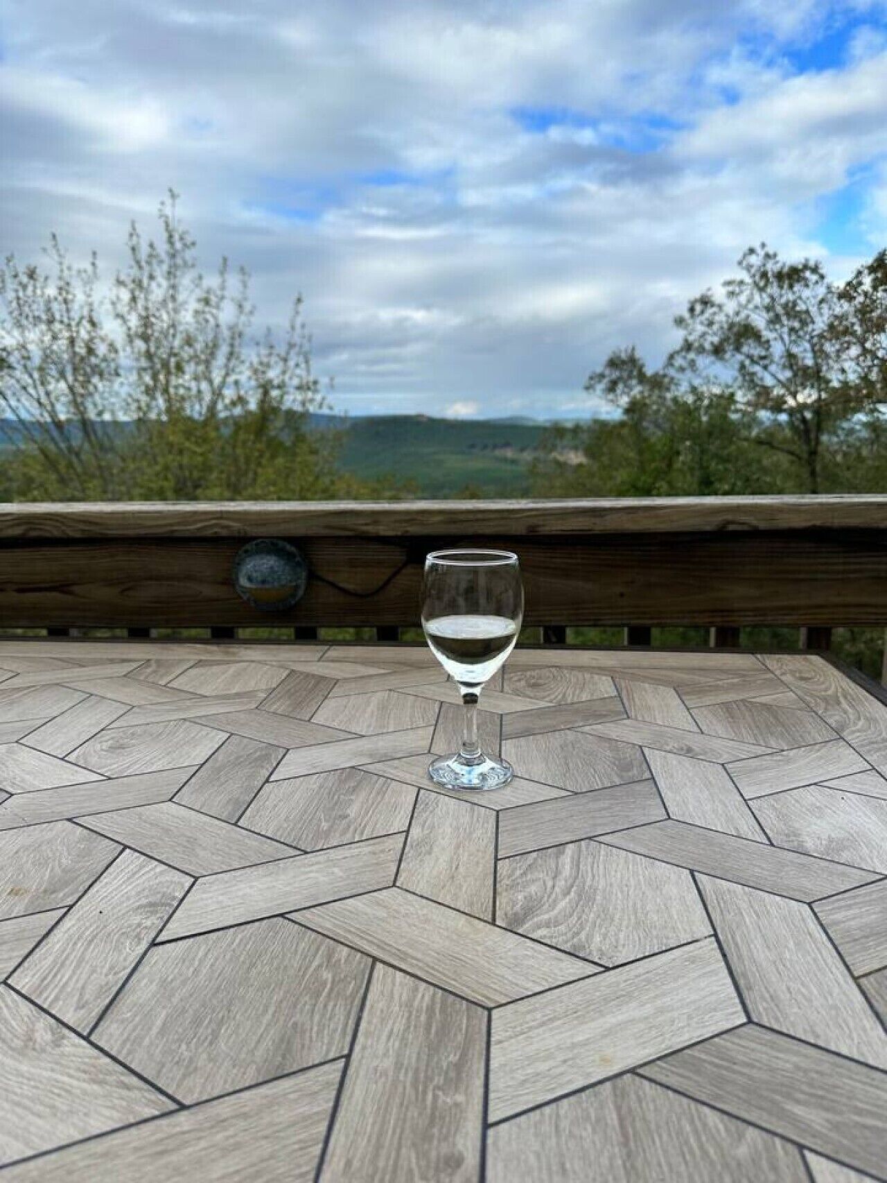 A glass of white wine on a wooden table with a geometric pattern, overlooking a scenic landscape with trees and hills.