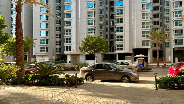 A modern high-rise residential building with multiple floors, a parking area with cars, and palm trees. - Prime Select Kattameya Bavaria (Cairo)
