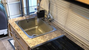 A compact kitchen with a stainless steel sink, a black stovetop with four knobs, and a small window above the sink. - Road Surfer-- Resort-Style RV Stay Near The Strip (Las Vegas)