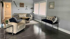 Un salon moderne avec un canapé d’angle, une table basse en verre et une table d’appoint en métal. Il y a deux fenêtres équipées de stores, une cheminée et une œuvre d’art accrochée au mur.