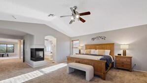Une chambre à coucher avec un grand lit, une tête de lit en bois, une table de chevet, un banc, un ventilateur de plafond et une cheminée.
