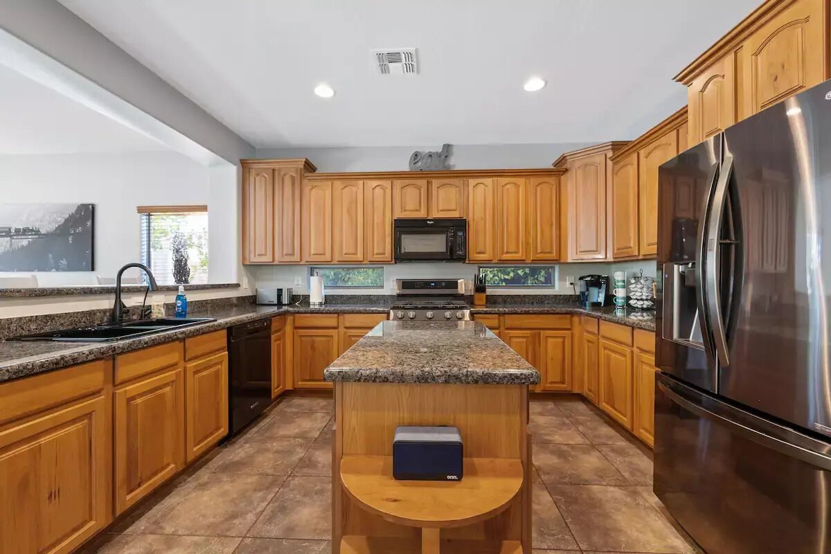 A modern kitchen with wooden cabinets, granite countertops, a stainless steel refrigerator, and a central island with a built-in oven.