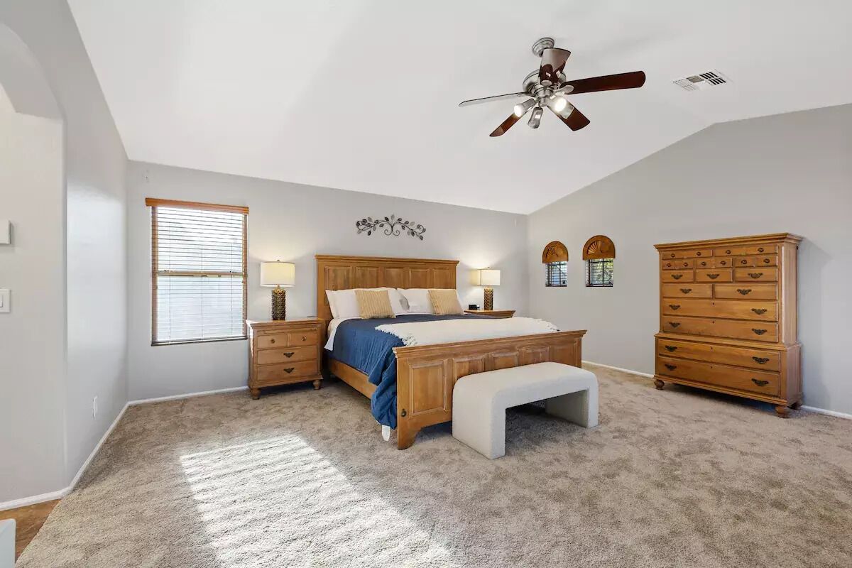 Une chambre à coucher comprenant un lit, des tables de chevet, une commode, un ventilateur de plafond et une fenêtre avec des stores.