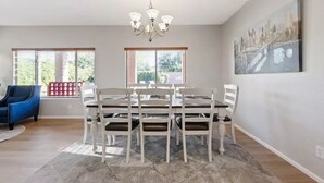 Un coin repas avec une table et des chaises blanches, un lustre et de grandes fenêtres équipées de stores.
