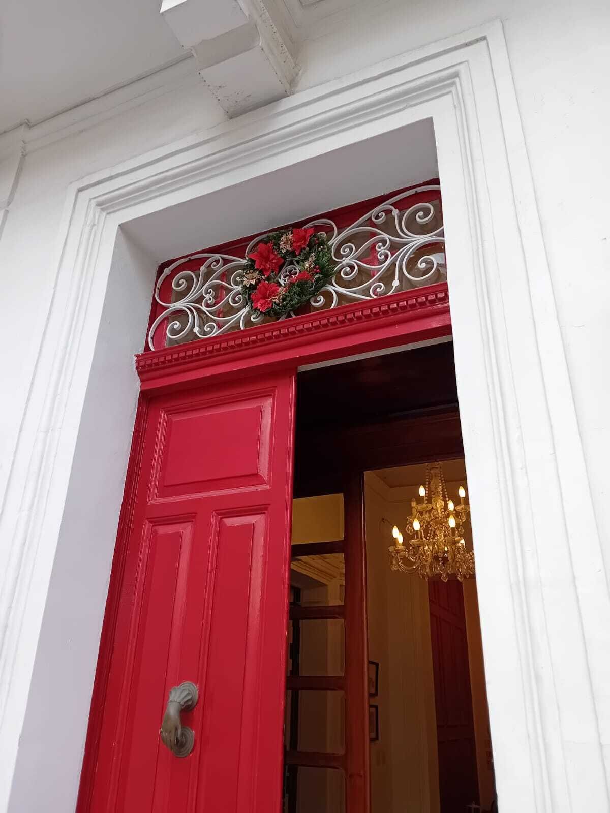 Une porte rouge ornée d’une guirlande décorative, et on peut apercevoir un lustre à l’intérieur.