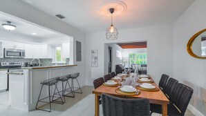 A modern kitchen with white cabinets, a microwave, and a dining area with a wooden table set for six. - Upper East Side - Miami Home - We Host (Miami)