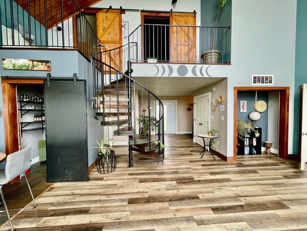 Un interior amplio con una escalera de madera que lleva a una zona superior tipo loft, un comedor con mesa y sillas, y un pasillo con una puerta y una pequeña mesa con plantas.