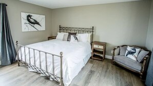 Un dormitorio con estructura de cama metálica, mesita de noche de madera, sofá gris y un cuadro de un pájaro colgado en la pared.