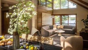 A spacious living room with a high ceiling, a dining area with a table set for a meal, and a balcony with a view of the outdoors. - TC Lake House w/Hot Tub, Pontoon, & Game Room! (Traverse City)