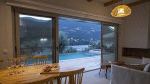 A dining area with a wooden table and chairs, a sofa, and a view of a mountain and pool. - Sea View Villa Selina with private sea access and infinity pool (Sivota Lefkada)
