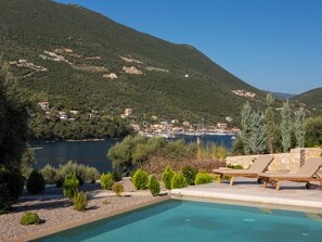 A pool with lounge chairs, surrounded by greenery and overlooking a lake with boats and buildings in the background. - Sea View Villa Anasa with private sea access and infinity pool (Sivota Lefkada)