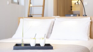 Una cama bien tendida con sábanas blancas, una bandeja con dos vasos de una bebida blanca y un pequeño jarrón con una planta verde, y una escalera de madera apoyada contra una pared al fondo.