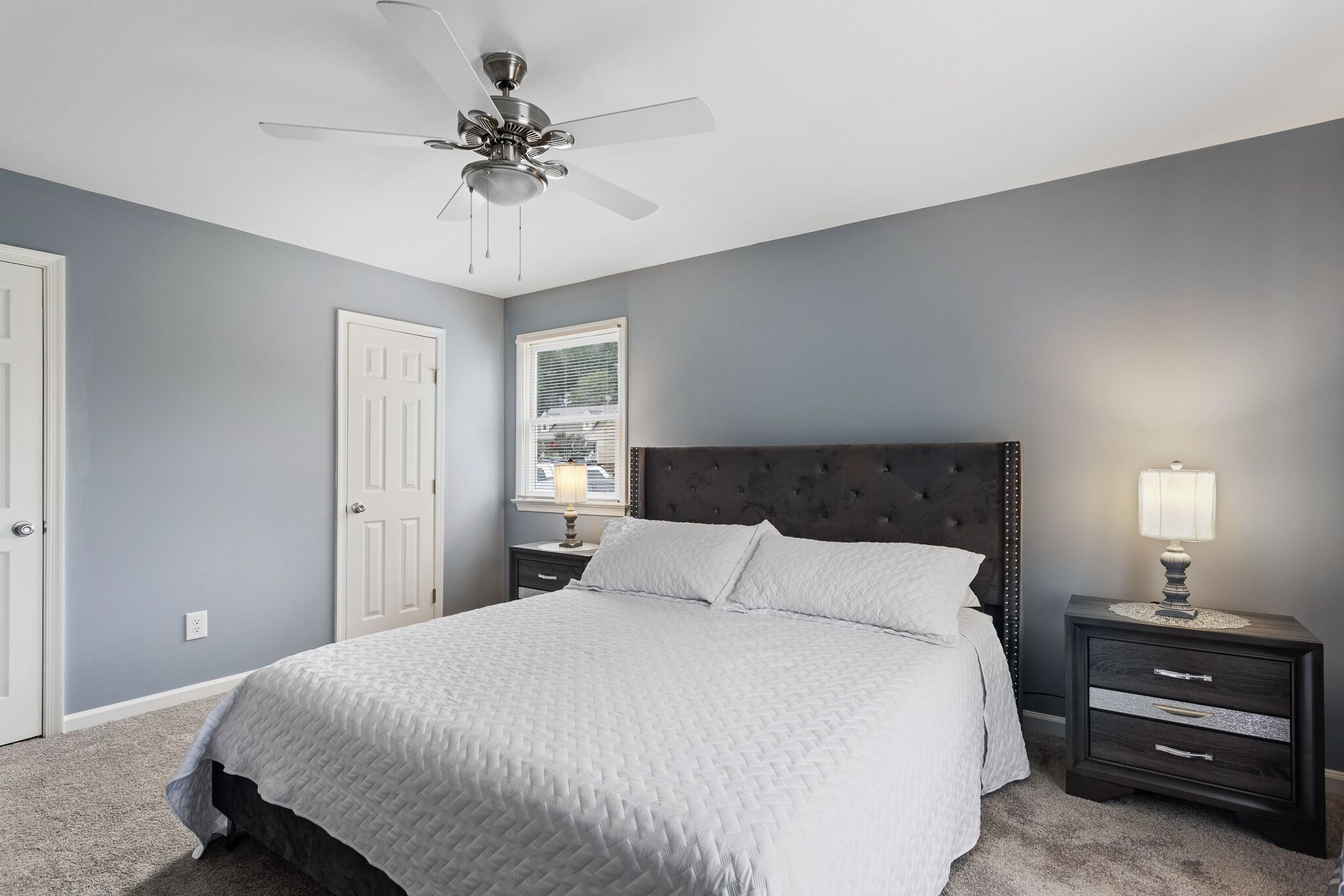 A bedroom with a bed, a nightstand with a lamp, a ceiling fan, and a door.