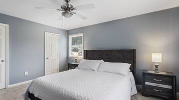 A bedroom with a bed, a nightstand with a lamp, a ceiling fan, and a door.