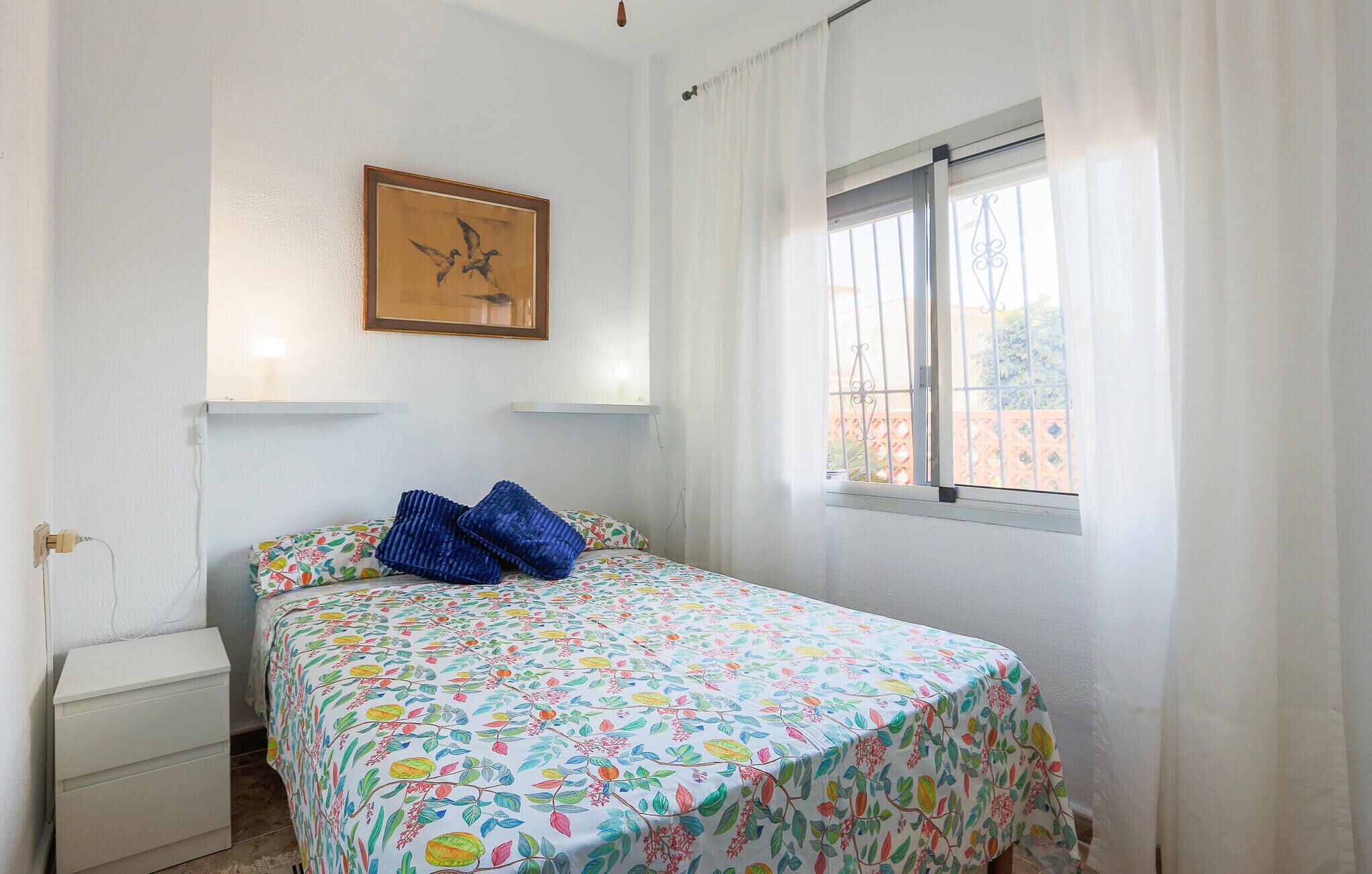 A bedroom with a bed, a nightstand, a window with curtains, and a wall-mounted painting.