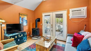 Una sala de estar acogedora con un televisor sobre una maleta antigua, una chimenea, un sofá con cojines de colores y un ventanal con vistas a muebles de exterior.