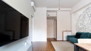 A modern living room with a flat-screen TV, a wooden floor, a green sofa, and a desk with a chair. - GuestHost - Modern and welcoming 80m2 apartment, ideal for 4 people, located on the mezzanine floor of a building (NO lift) in the Bullona area. The property is located in a strategic area, a few minutes walk from the “Gerusalemme” and “Domodossola (Milano)