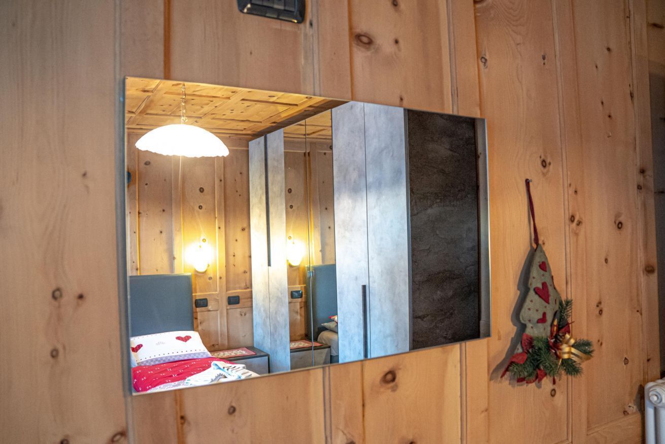 A wooden room with a bed, bedside table, and a mirror reflecting the room.
