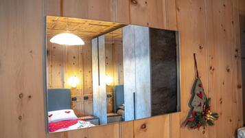 A wooden room with a bed, bedside table, and a mirror reflecting the room.