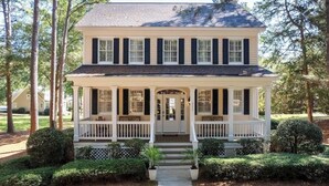 Une maison à deux étages avec un porche, entourée d’arbres et agrémentée d’un aménagement paysager soigné.
