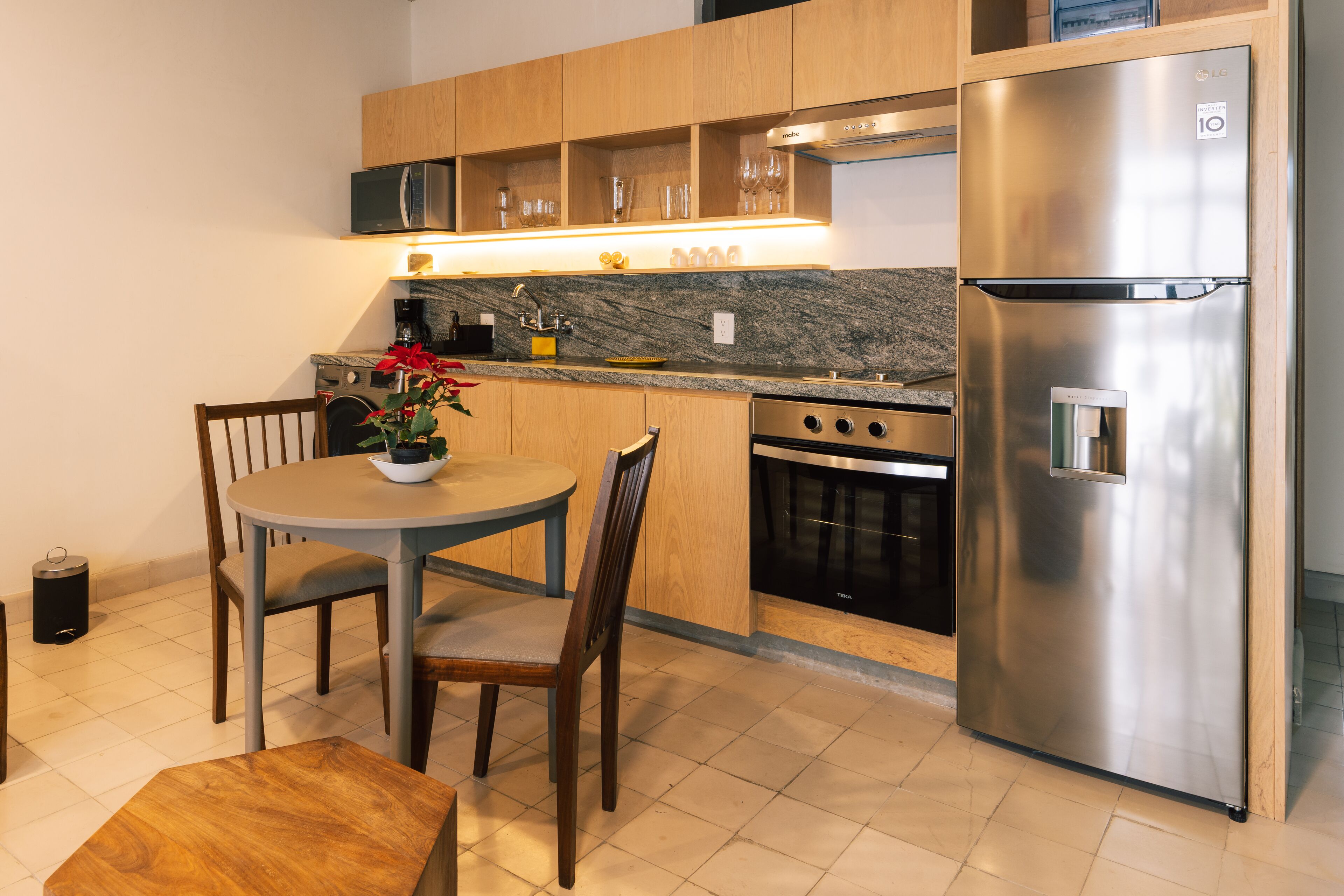 Una cocina compacta con armarios de madera, un refrigerador de acero inoxidable, un horno empotrado y un comedor con una mesa redonda y sillas.