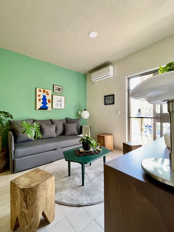 Una sala de estar moderna con una pared verde, un sofá gris, una mesa de centro con una planta y un ventanal que proporciona luz natural.