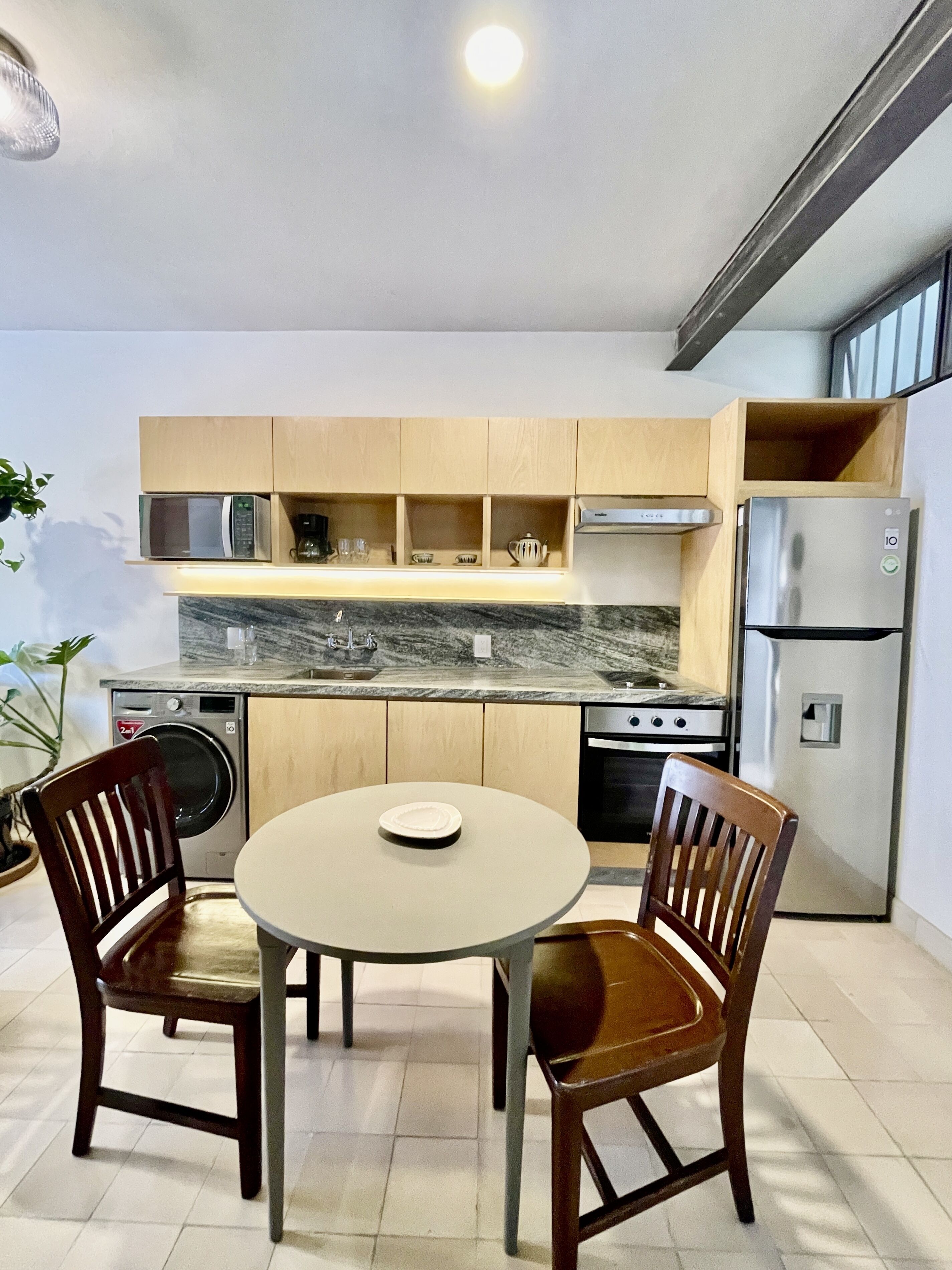 Een kleine keuken met houten kastjes, een ingebouwde magnetron, een roestvrijstalen koelkast en een ronde tafel met twee stoelen.