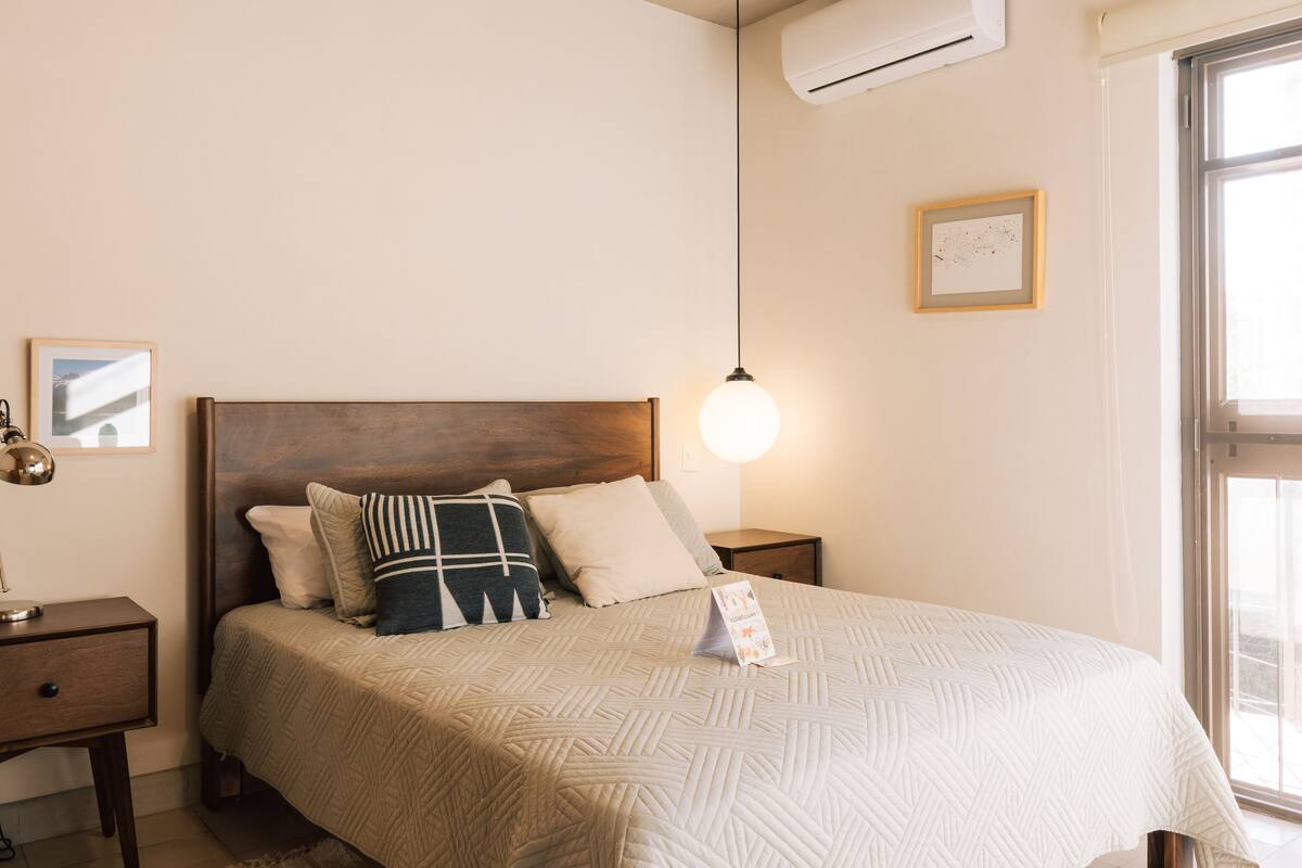 Een slaapkamer met een bed, nachtkastjes, een vloerkleed, een airconditioner en een raam.