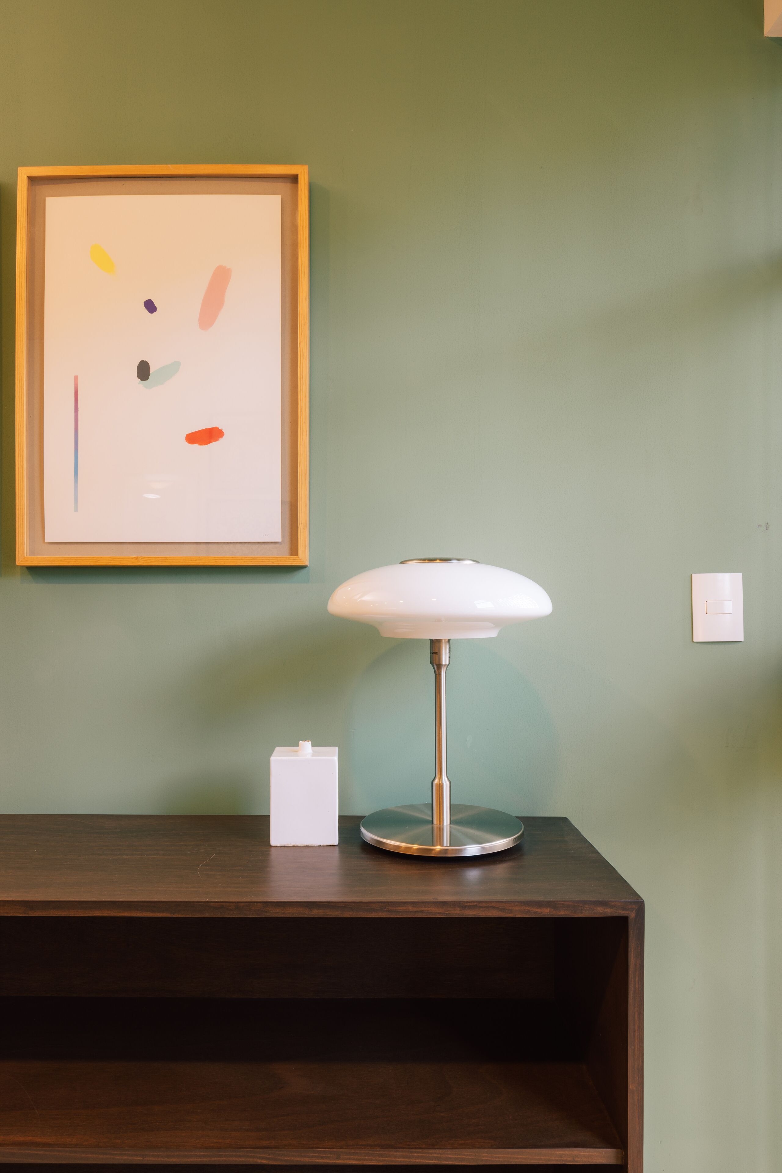 Un cuadro abstracto enmarco, una lámpara blanca y una mesa de madera oscura frente a una pared verde.