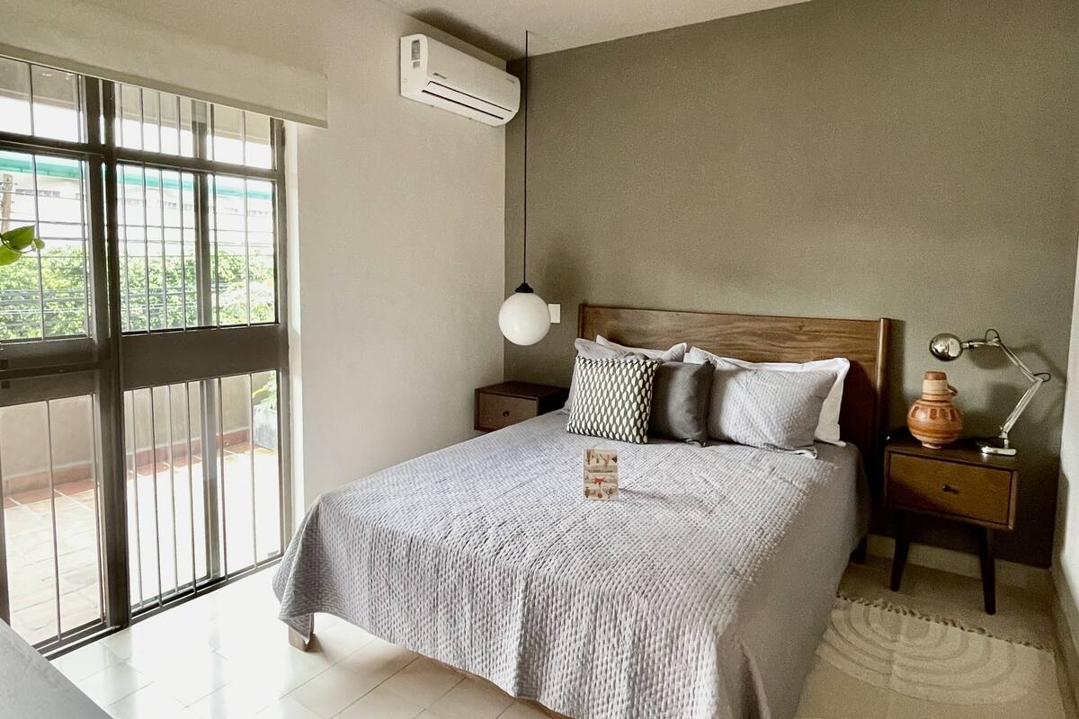 Een slaapkamer met een bed, nachtkastjes, een lamp, een airconditioner en uitzicht door een raam.