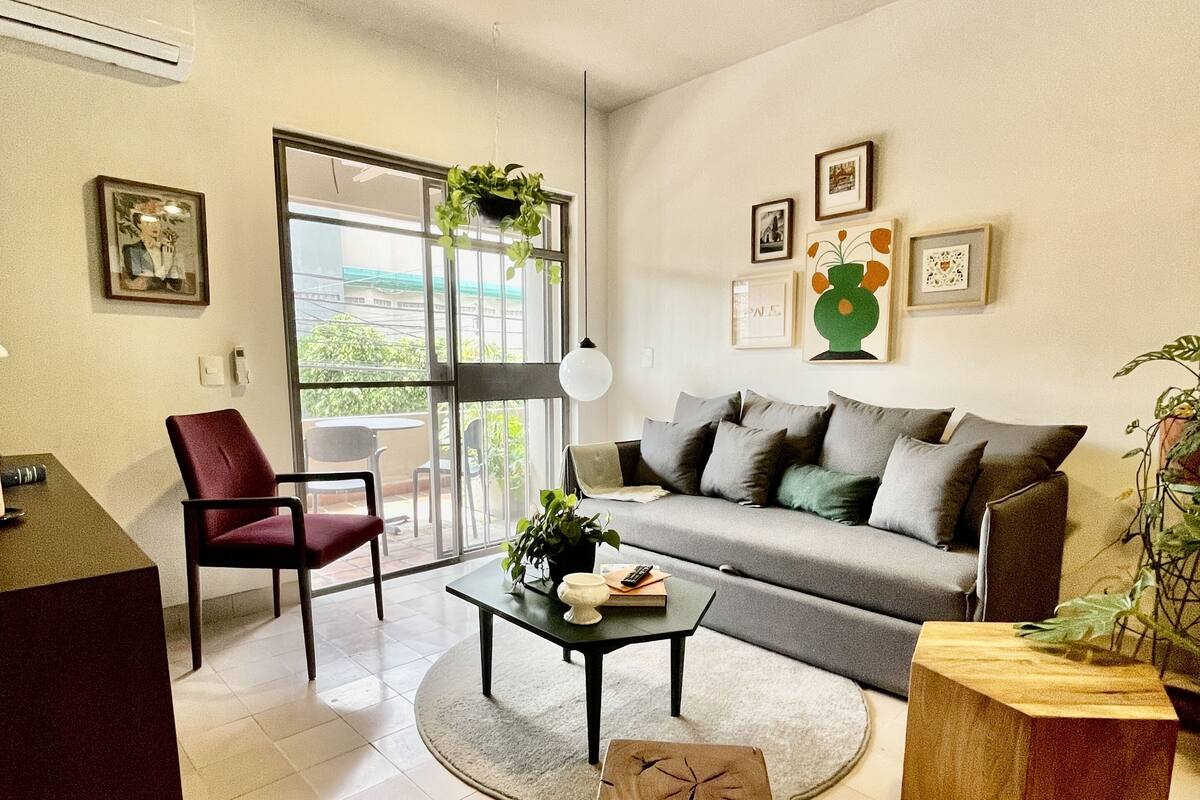 Een moderne woonkamer met een bank, een salontafel, een rode fauteuil, een houten bijzettafeltje en een klein krukje. Er is een raam met uitzicht op groen.