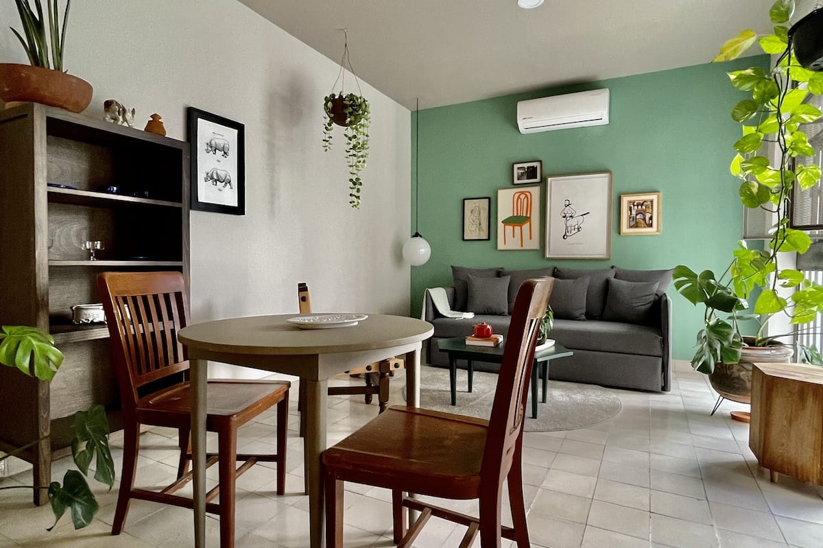 Een moderne woonkamer met een eetgedeelte, met een ronde tafel, houten stoelen, een bank en een salontafel. Er staan diverse potplanten en er hangen afbeeldingen in lijsten aan de muren.