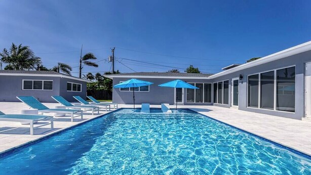 A swimming pool with lounge chairs and umbrellas, surrounded by a modern building with large windows. - 2025-C Sunshine Manors (Wilton Manors)