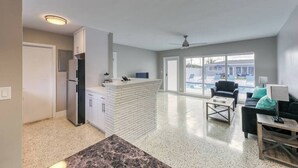 A spacious living area with a kitchen, a dining table, and a large window offering a view of a pool and outdoor seating. - 2025-C Sunshine Manors (Wilton Manors)